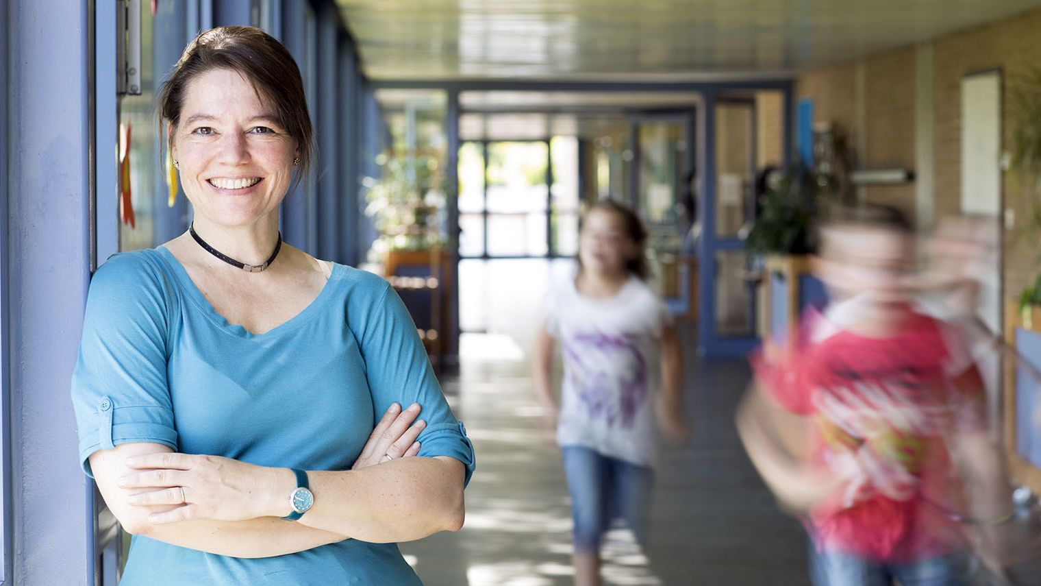 A female teacher stands in a school hallway, she smiles at the camera, her arms are crossed. Children are running in the background.