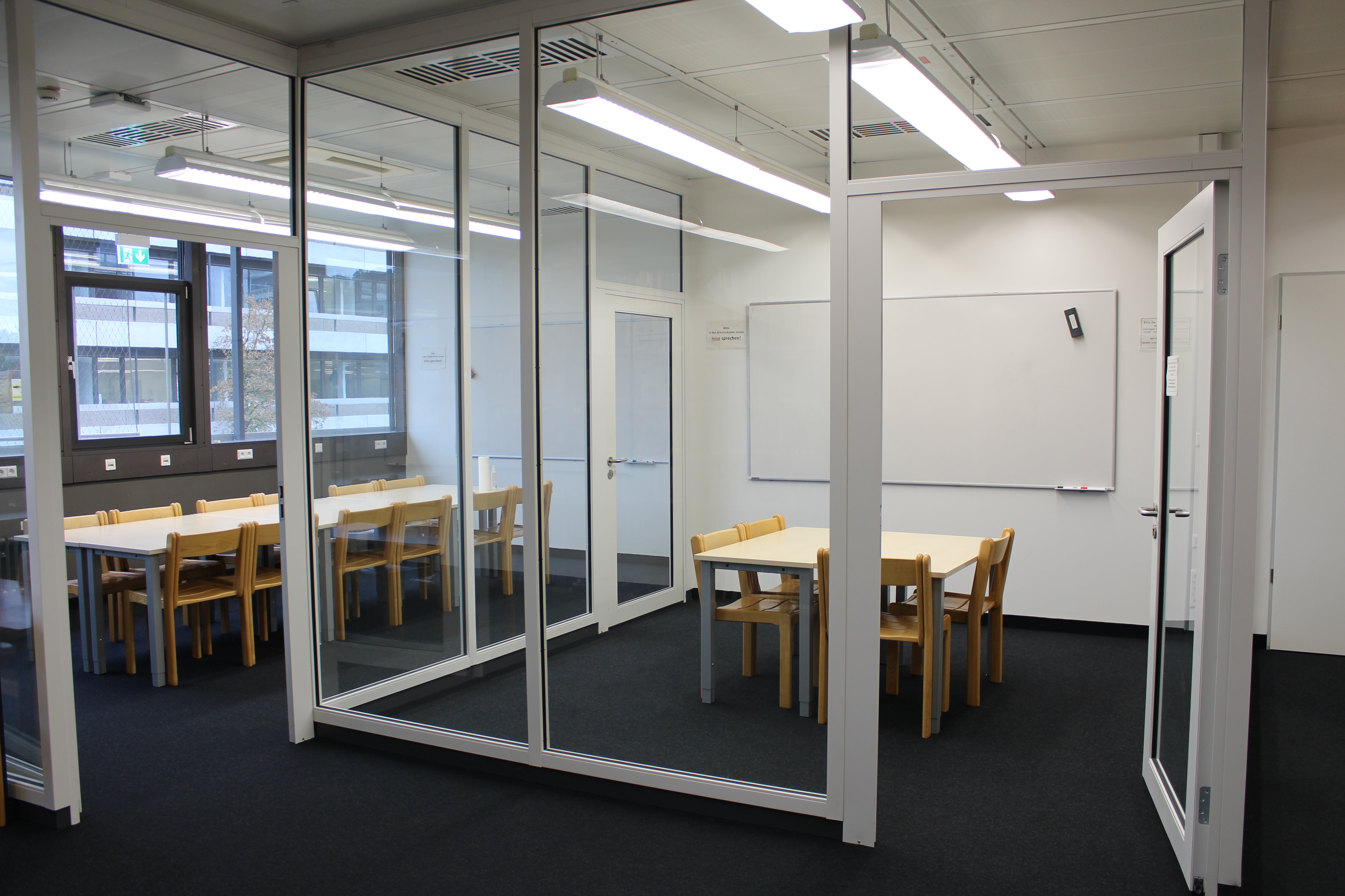 Two group rooms within the library are separated from the rest of the room by glass walls. There are tables, chairs and a whiteboard in each room.